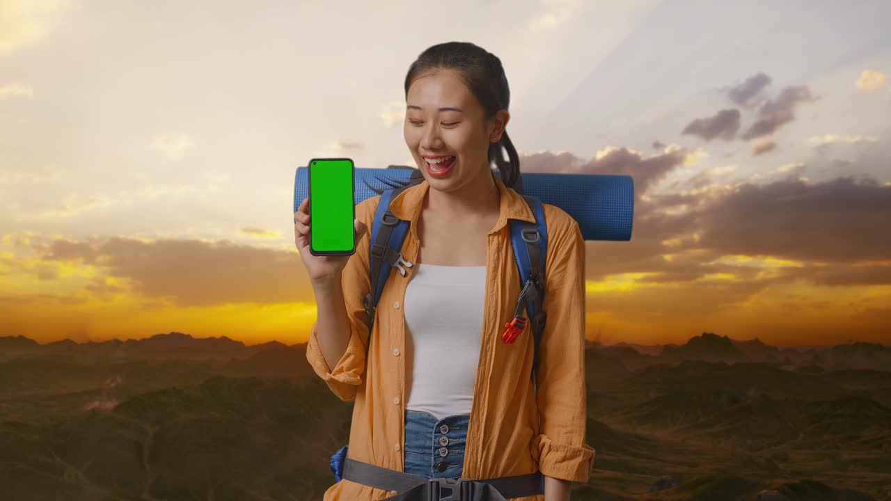 excursionista asiática con mochila de montañismo sonriendo y mostrando una maqueta de pantalla verde de un teléfono inteligente mientras está de pie en la cima de la montaña durante la hora del atardecer