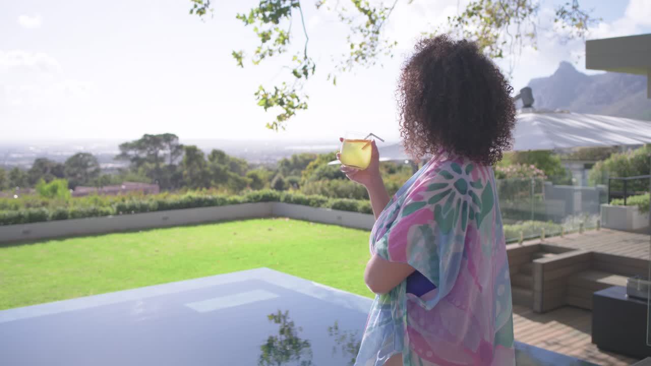 Donna in copricostume pastello attratta dal sole verso il bordo di una piscina a sfioro, solleva un bicchiere, sorseggia, con spazio per il testo