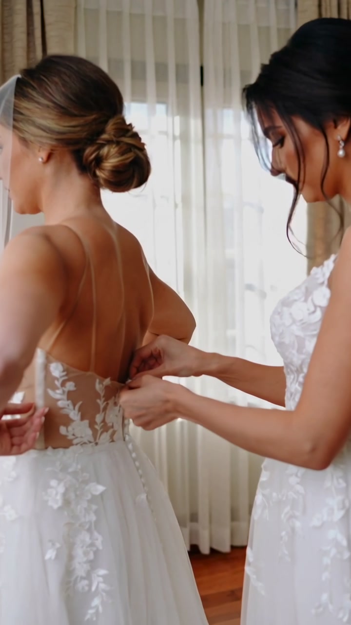 Vertical video: Reaching bride's back, helper fastening gown buttons to secure fit in dressing room