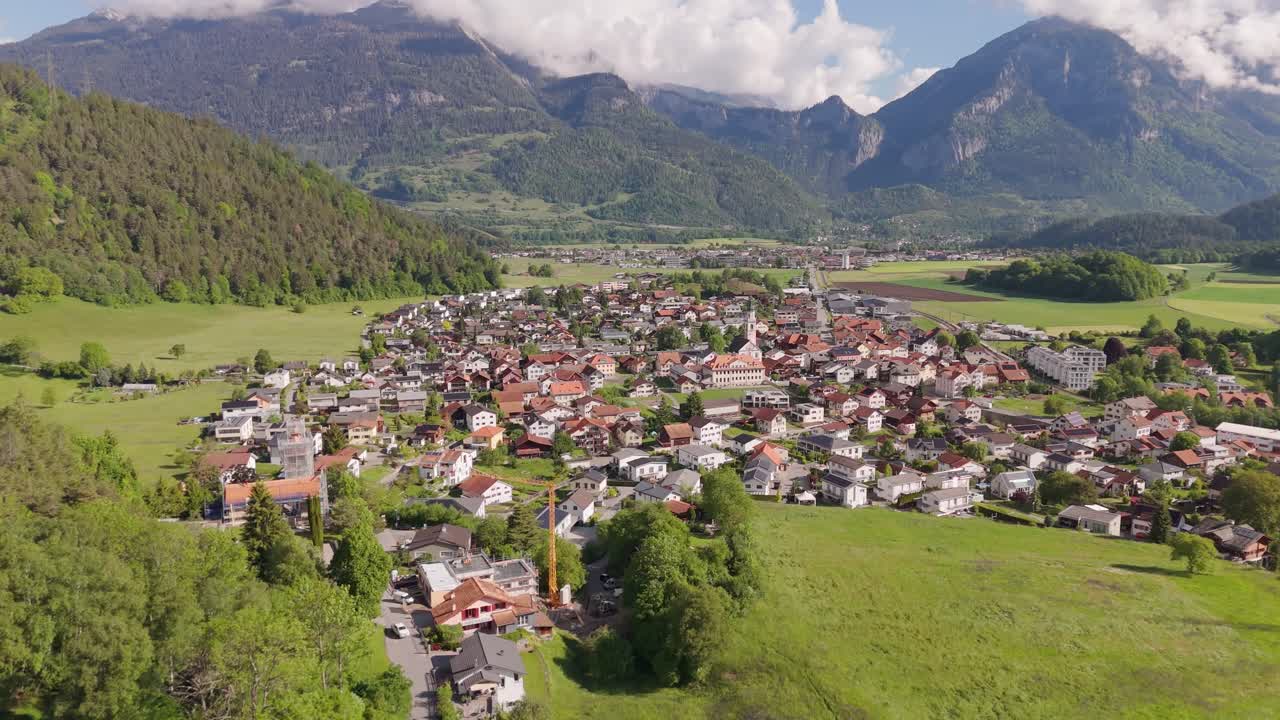 Aerial approaching shot of historic town of Rhäzüns, Switzerland during sunny day in spring. Green forest trees on hillside. WIde shot. Alps mountains in distance. Quiet calm in swiss neighborhood.