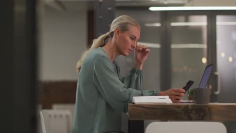 caucasian businesswoman sitting at desk using laptop and smartphone wondering