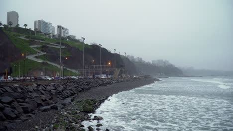 Static-shot-of-a-foggy-morning-along-the-coast-of-Punta-Roquitas,-Malecon-de-Miraflores,-Lima,-Peru,-with-waves-crashing-onto-the-rocky-beach