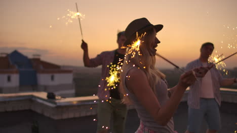 the girl in the hat beautifully moves her hands in a dance with big bengal light. she is on the party with her friends on the roof.