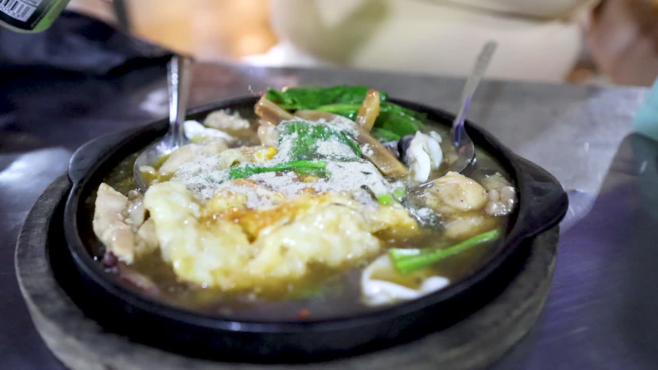 Hand pours seasoning onto sizzling noodle dish in vibrant Bangkok street food market, close-up view