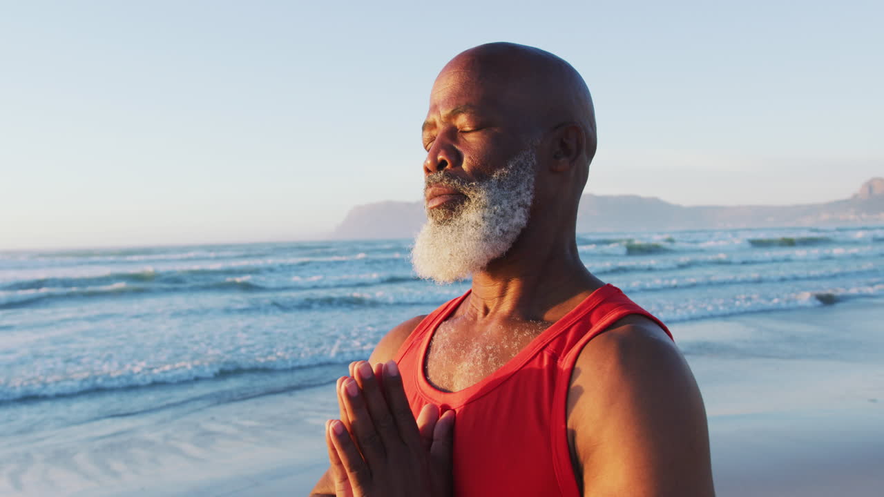 un anciano afroamericano practicando yoga con los ojos cerrados en la playa.
