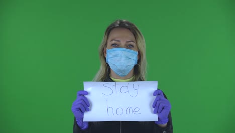 portrait of beautiful young woman in medical mask is looking at camera and holding a poster with the words stay home. blonde with loose hair in a black jacket and jeans on a green screen in the studio. health protection corona virus concept