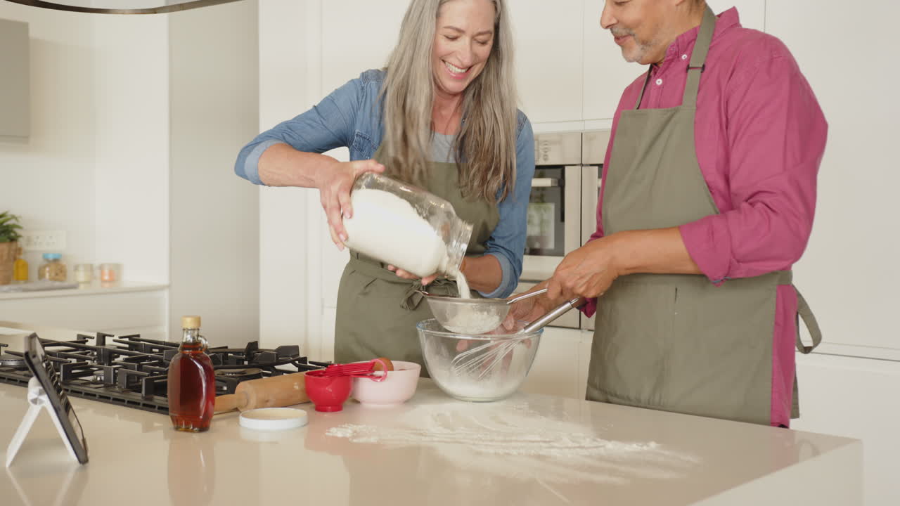 Baking together, multiracial senior couple sifting flour into bowl in modern kitchen, at home