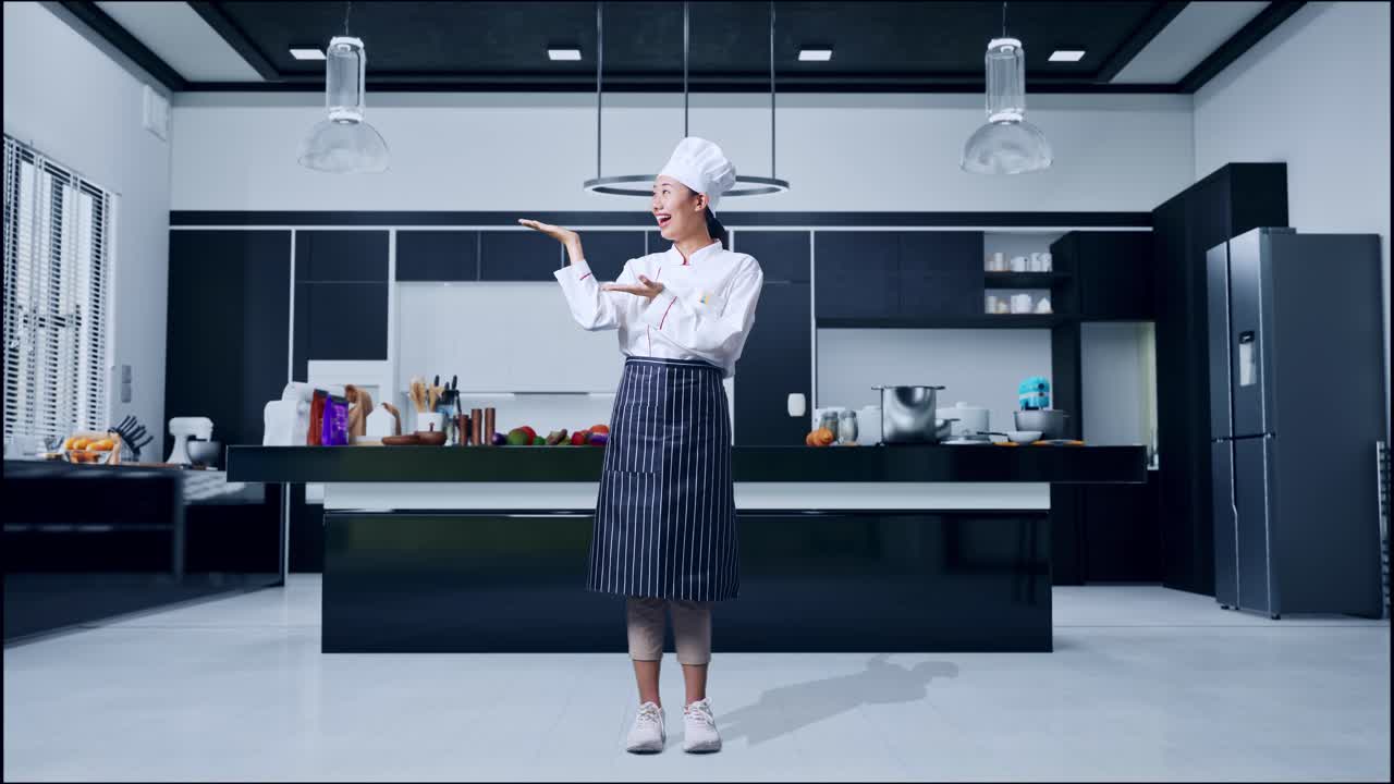 Figura intera di una chef asiatica che sorride e indica di lato nella cucina di casa.