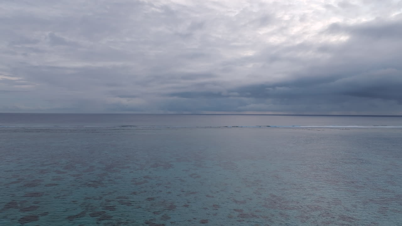 Aerial view of clear ocean water with coral reefs visible beneath the surface. Captured by a drone moving forward over the sea under an overcast sky in the Cook Islands