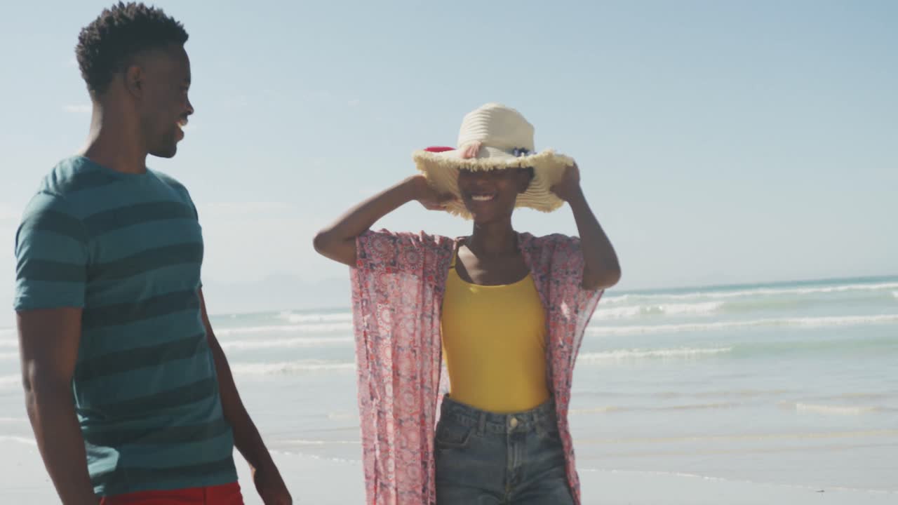 Happy african american couple walking with hat on sunny beach