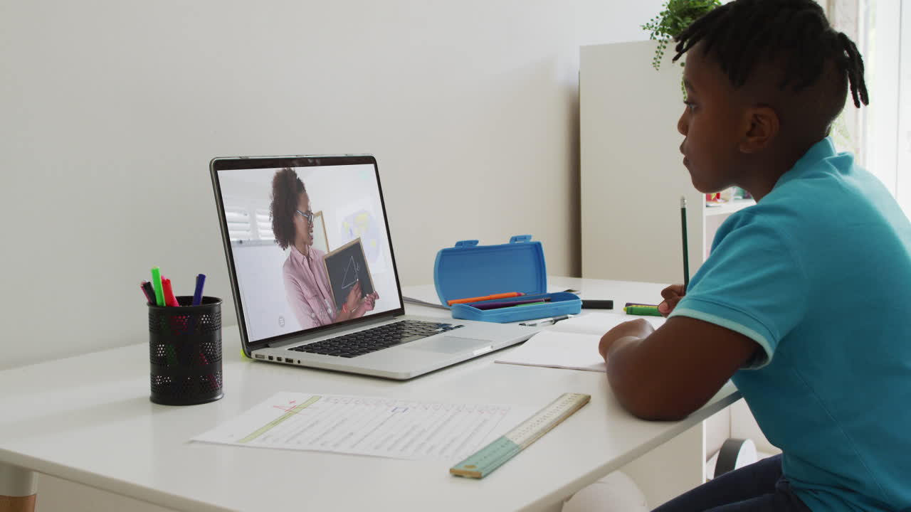 niño afroamericano haciendo la tarea mientras tiene una videollamada con una maestra en una computadora portátil en casa