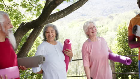 happy diverse senior male and female friends holding yoga mats talking in sunny nature, slow motion