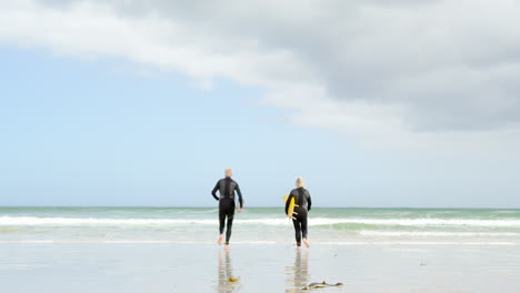 Rückansicht-Eines-Alten-Kaukasischen-Seniorenpaares,-Das-Mit-Surfbrett-Am-Strand-Läuft-4k