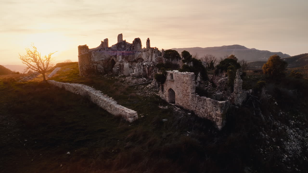 Ripresa aerea cinematografica 4K con drone che sorvola antiche rovine in pietra al tramonto, con una calda luce dell'ora d'oro su un paesaggio montano suggestivo. Mura storiche di una fortezza abbandonata in una campagna panoramica.