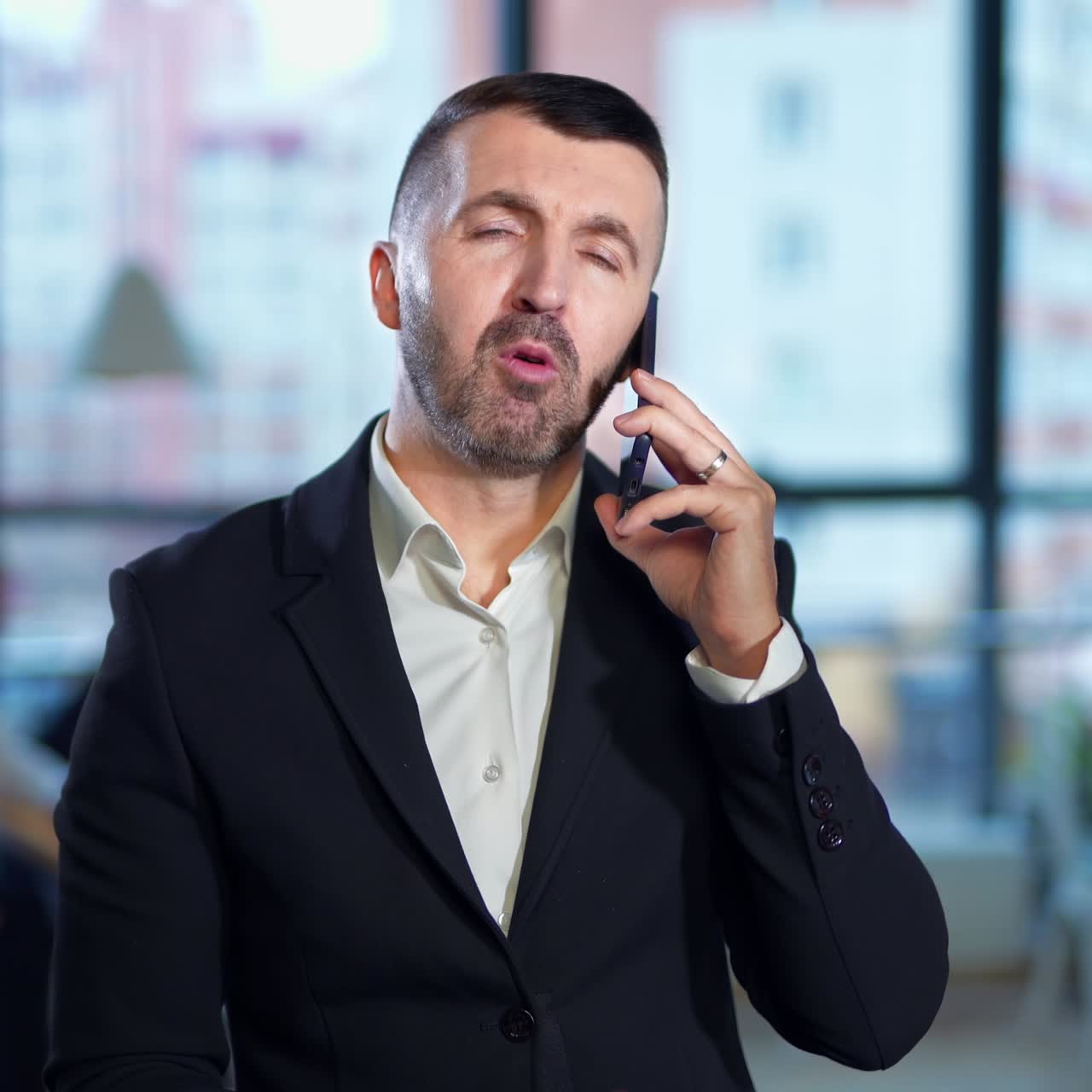 Usmívající se pozitivní vousatý muž hovoří po telefonu a mává rukou. Během hovoru se podnikatel dívá na hodinky. Kolegové sedí v pozadí u stolu, rozmazaní.
