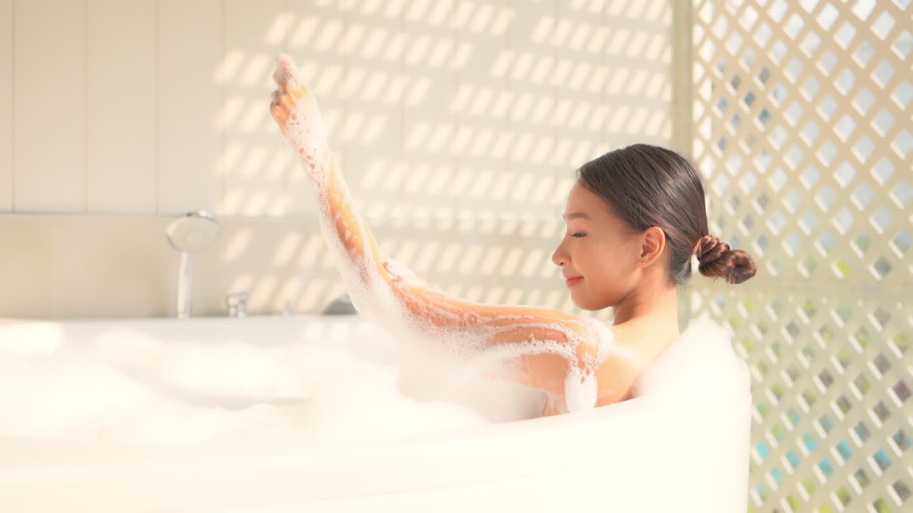 Static slow-motion shot of a sexy Asian lady relaxing in a bubble bath in a white tub and playing with soap