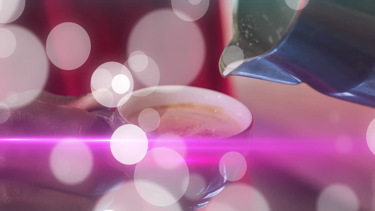 coffee shop barista hand gripping cup handle pouring coffee from stainless pitcher with pink bokeh