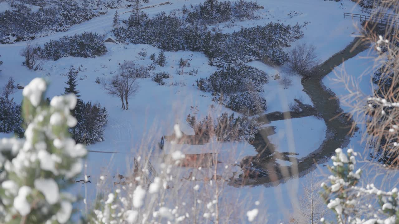 A small wooden cabin sits near a winding river in a snowy alpine valley surrounded by winter bushes.