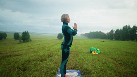 side view of a woman in a green black suit standing on a colorful mat, bringing her hands together slowly as she bows her head slightly while practicing yoga in a vast open field under a cloudy sky