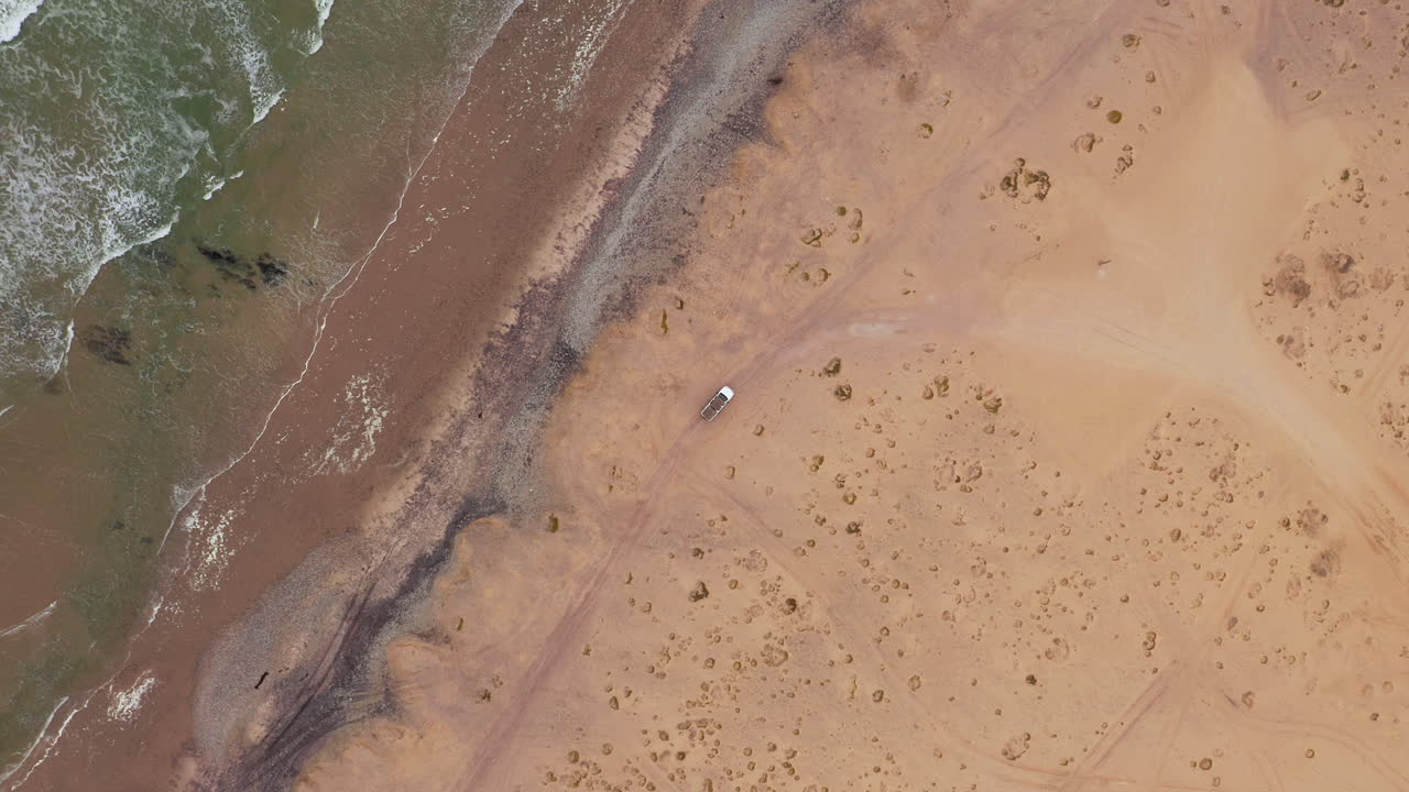 Top down drone shot of an off-road vehicle parked alone on a wide sandy beach near Swakopmund in Namibia, with soft waves rolling in under cloudy skies