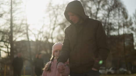 a little girl in a pink cap and jacket, and a man in a brown jacket, both wearing blue jeans, are holding hands and walking together in a park on a sunny day