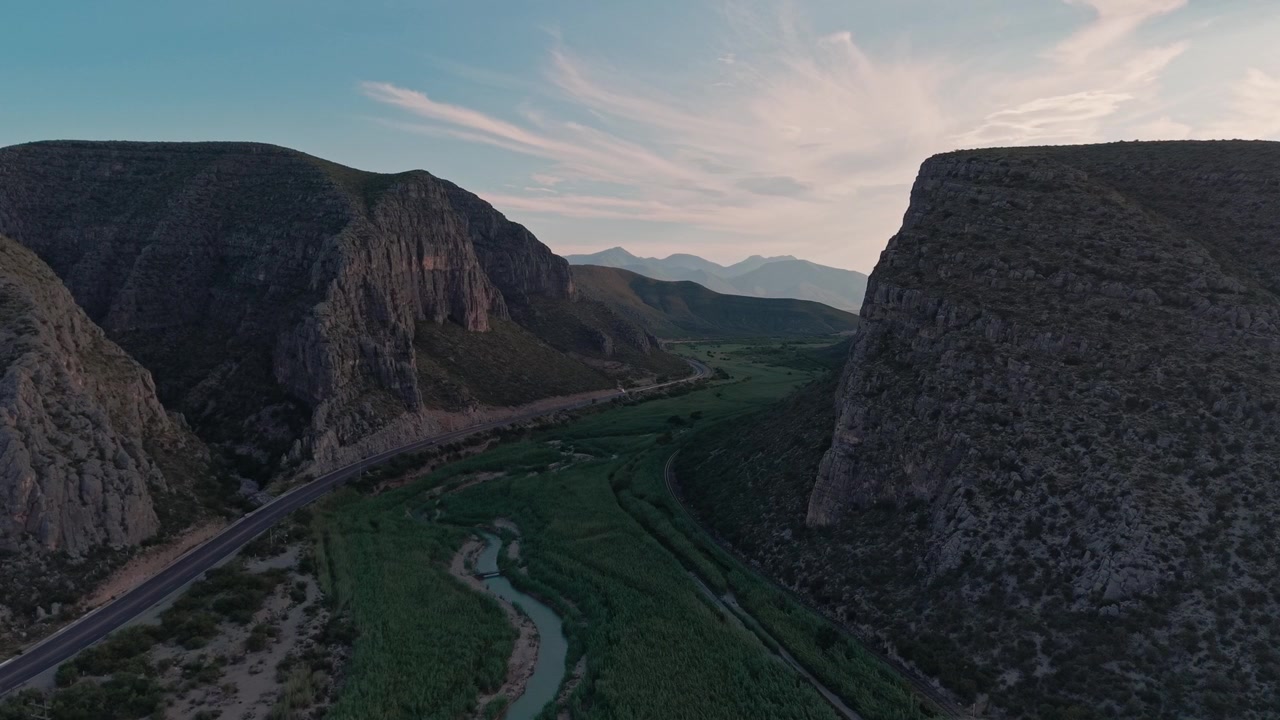 Aerial drone view of rugged canyon surrounding winding river and mountain road, with green vegetation and distant peaks beneath a partly cloudy evening sky