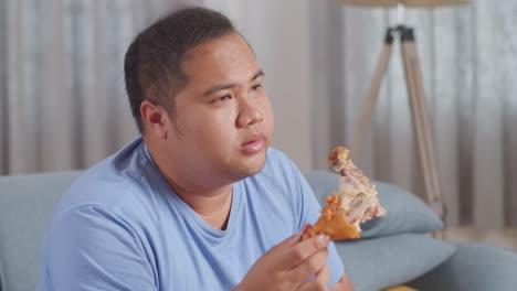 close up of a fat asian man having a headache while having fried chicken eating fast food on a sofa in the living room at home