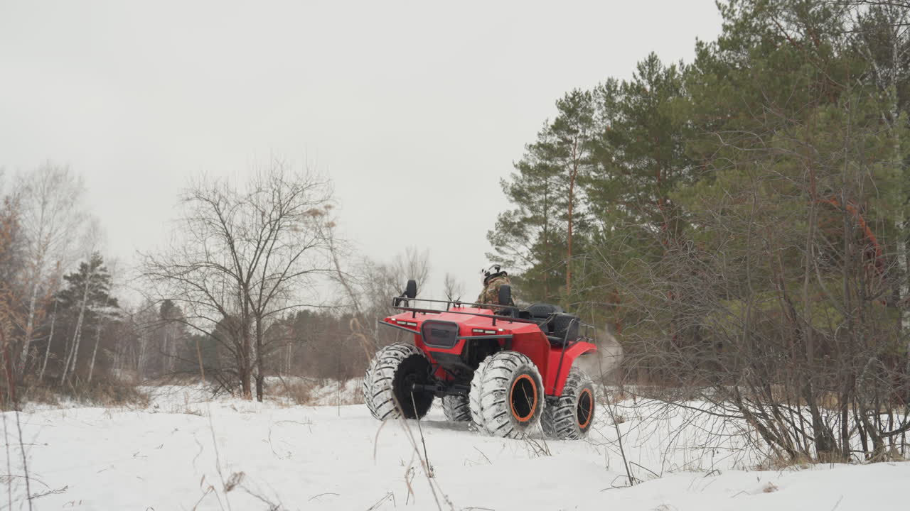 ATV rosso che sfreccia attraverso un campo innevato, pilota con casco e giacca invernale che traccia solchi lungo il bordo della pineta, neve sollevata dagli pneumatici sovradimensionati, cielo coperto, fuoristrada estremo e adrenalinico