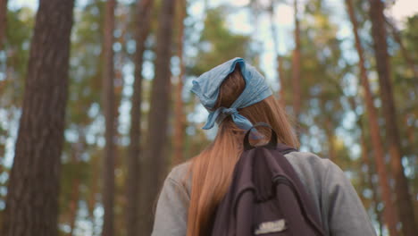 back view of young woman hiking through vibrant forest, wearing blue bandana and backpack, looking upward amidst tall trees, sunlight filters through foliage