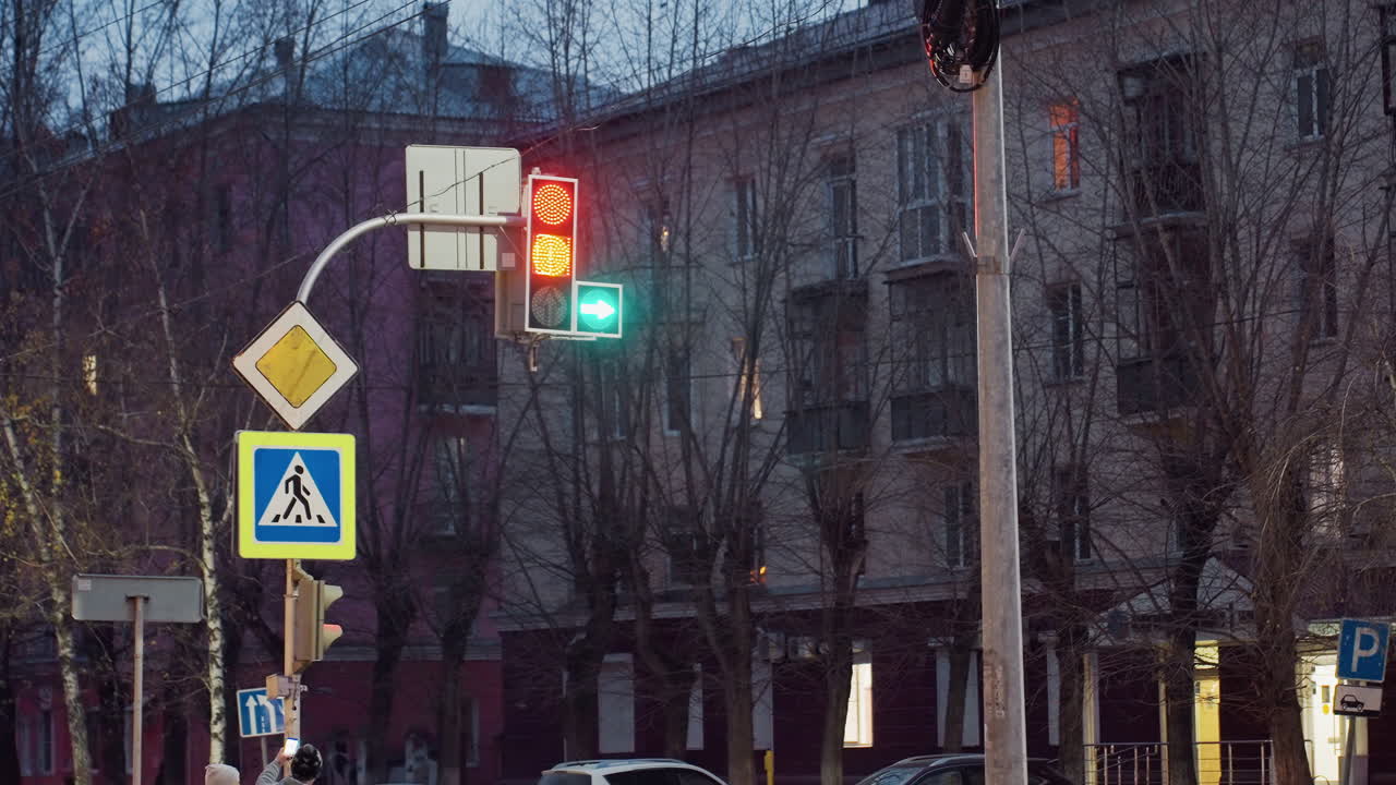 Urban intersection scene showing traffic light displaying red stop and green arrow signal surrounded by road signs, leafless trees and residential buildings in twilight city atmosphere