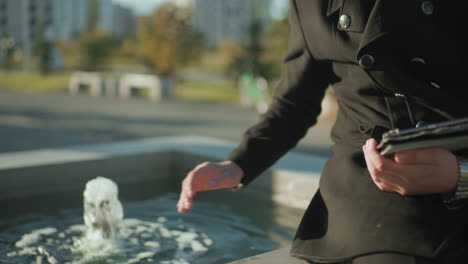 close up of man in black coat multitasking with tablet near spring water outdoors, hand poised for interaction, bright sunlight illuminating scene with blurred urban greenery