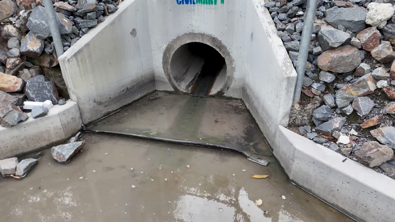 Stormwater slowly flows from concrete drainage culvert, surrounded by rocks, overcast natural lighting, static shot