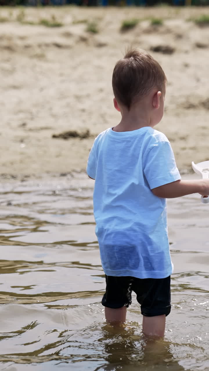 Adorable toddler boy standing in the water. Little kid plays with watering can and shovel on the beach. Vertical video
