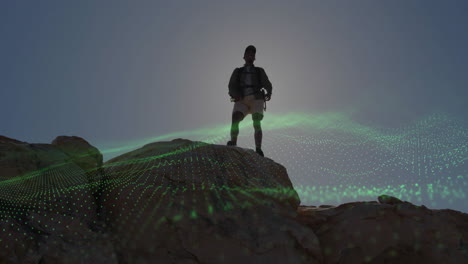 male hiker standing on rocky peak, showcasing green dotted digital wave overlay for technology