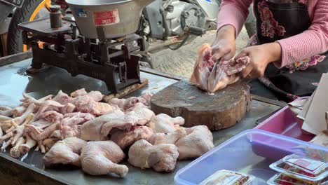 a traditional market vendor chopping raw chicken with a cleaver on a wooden block. fresh chicken meat is neatly arranged for sale beside an old-fashioned weighing scale,