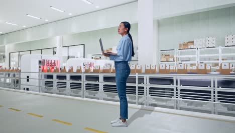 full body side view of asian female with conveyor for packaging coffee beans in bags at coffee factory, observes by looking up before she come to concentrating on the laptop and keep on typing