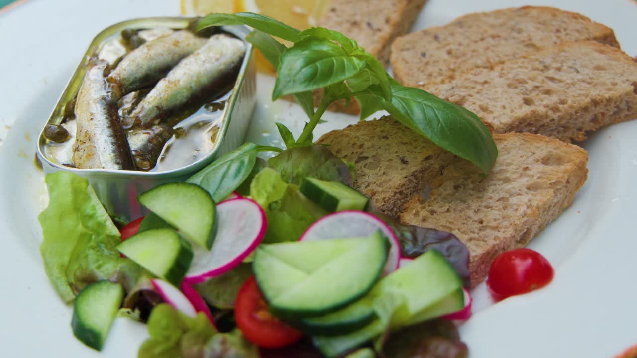 Hand serves sardines from tin onto plate with salad, bread, bright natural lighting, close-up