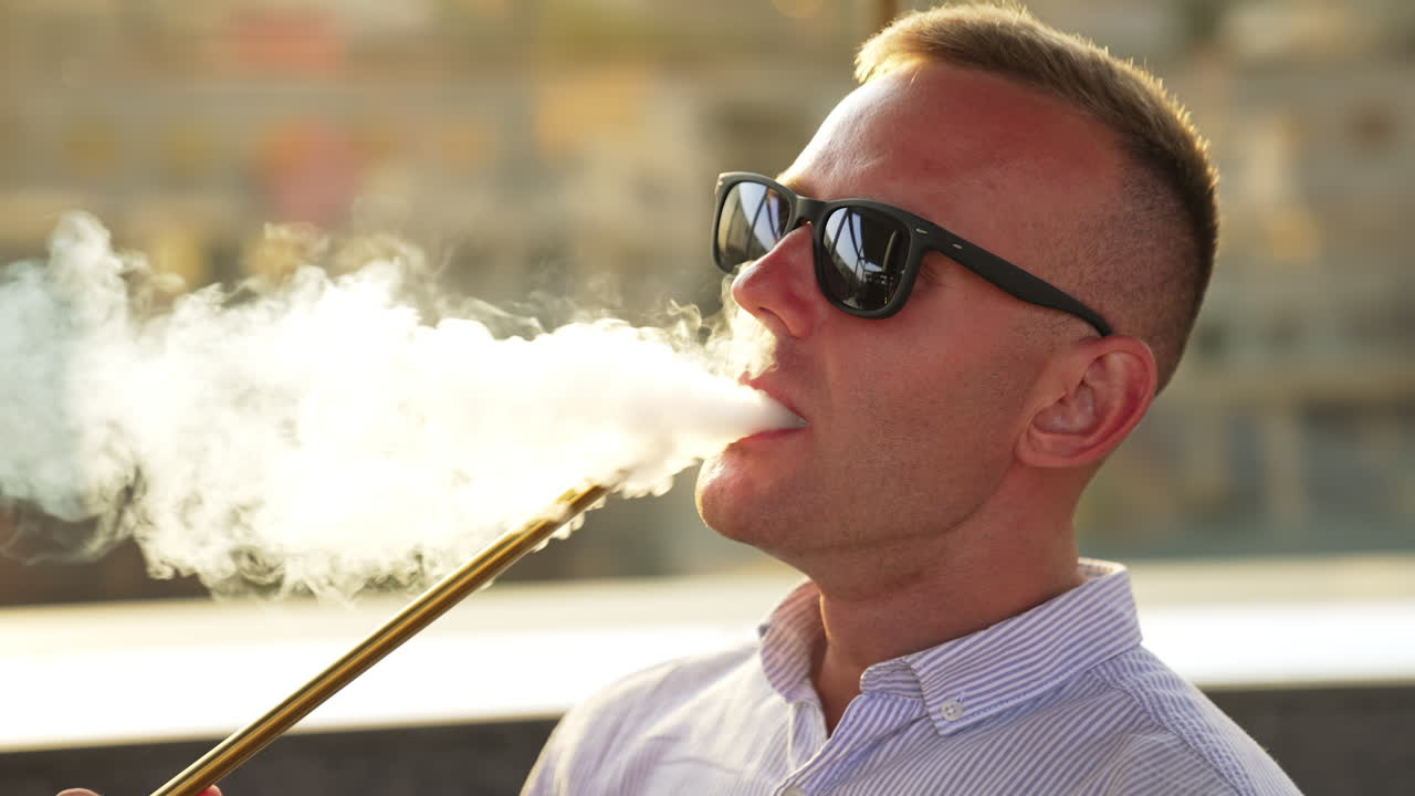Blond man wearing sunglasses smoking hookah. Man produces smoke from nose and mouth at once. Close up.