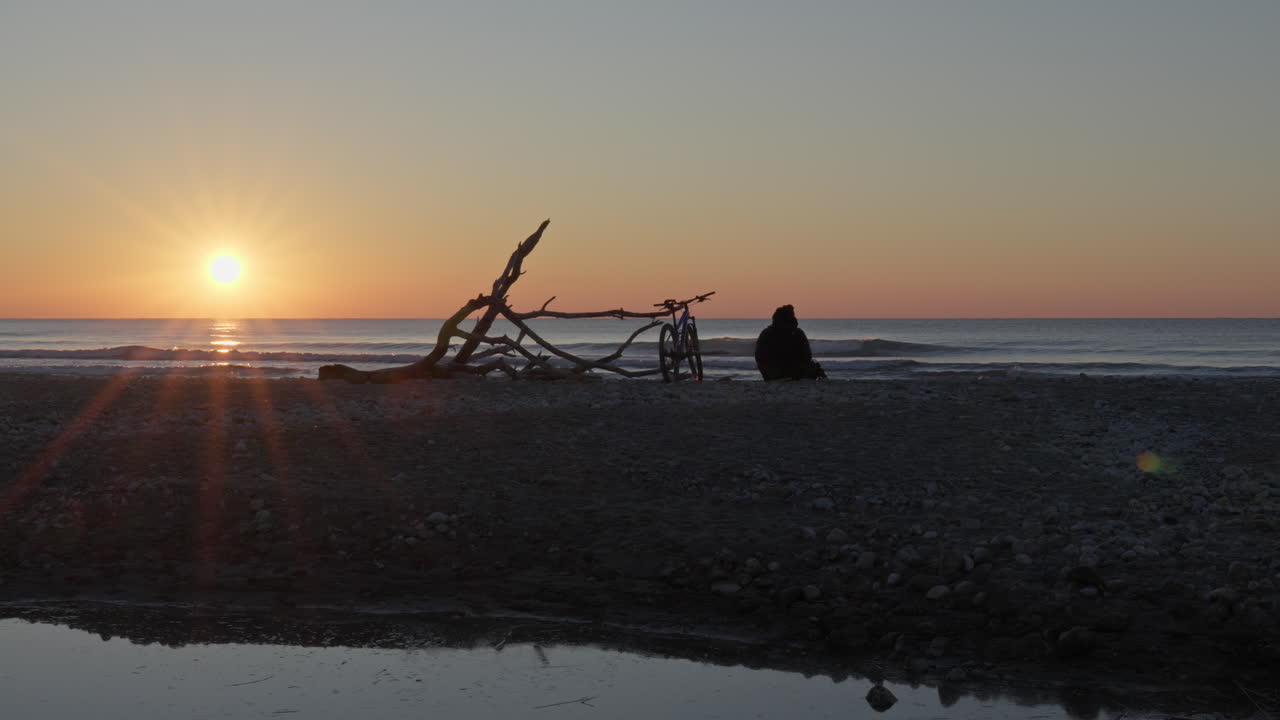 Time-lapse captures breathtaking beauty of a sunrise on beach, man arrives settles on ground near a fallen piece of driftwood, basked in the warm glow of the golden hour