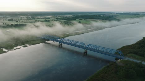 aerial establishing view of the steel bridge over lielupe river on a sunny summer morning, fog rising over the river, cars driving, wide drone shot moving forward