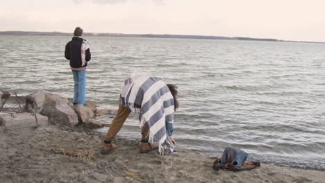 Rückansicht-Von-Zwei-Freunden-In-Winterkleidung,-Die-An-Einem-Bewölkten-Tag-Kieselsteine-An-Einer-Küste-Ins-Wasser-Werfen