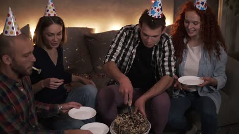 Birthday-guy-in-plaid-shirt-cutting-festive-birthday-cake-in-colorful-cone-hat.-Cheerful-friends-in-cones-enjoying-celebration-in-festive-decorated-room.-Sharing-plate-for-cake-and-smiling