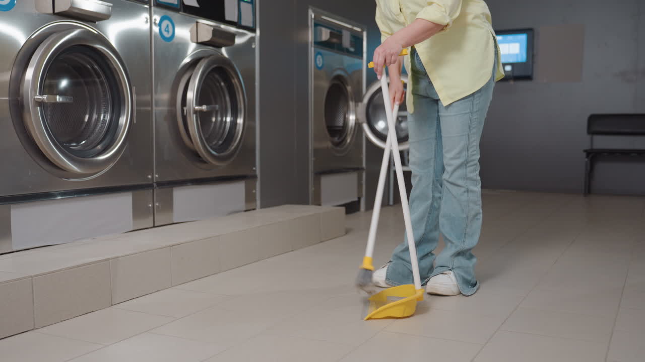 Personnel cleaning floor with broom and dustpan inside modern laundromat, stainless washers along wall, yellow shirt worker sweeping debris after filter drawer service, maintaining hygiene