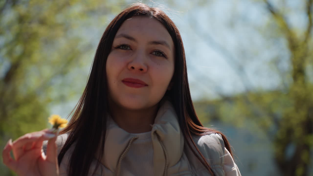 도시 공원에서 민들레를 들고 있는 여성이 배경에 건물을 두고 사색에 잠긴 표정과 편안한 자세를 취하고 있어요. 밝은 봄 햇살, 차분한 코트, 멀리 보이는 스카이라인이 도시와 자연의 대비를 만들어내고 있습니다.