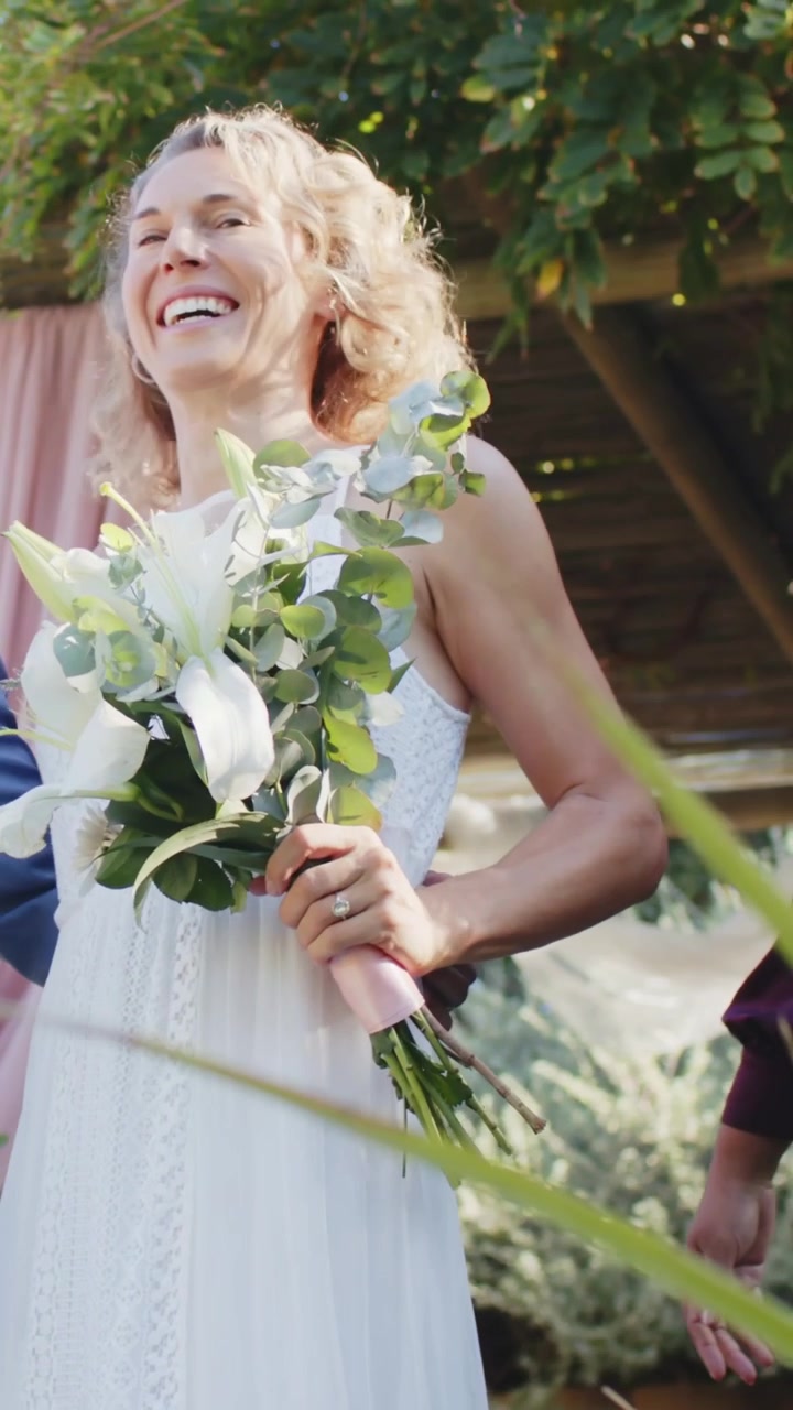 Vertical video: Laughing bride in white dress with bouquet in garden groom placing arm showing ring