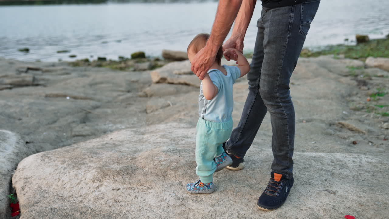Small kid stepping excitedly by the big smooth stones. Dad supporting little child by the hands. River and wooded bank at backdrop.