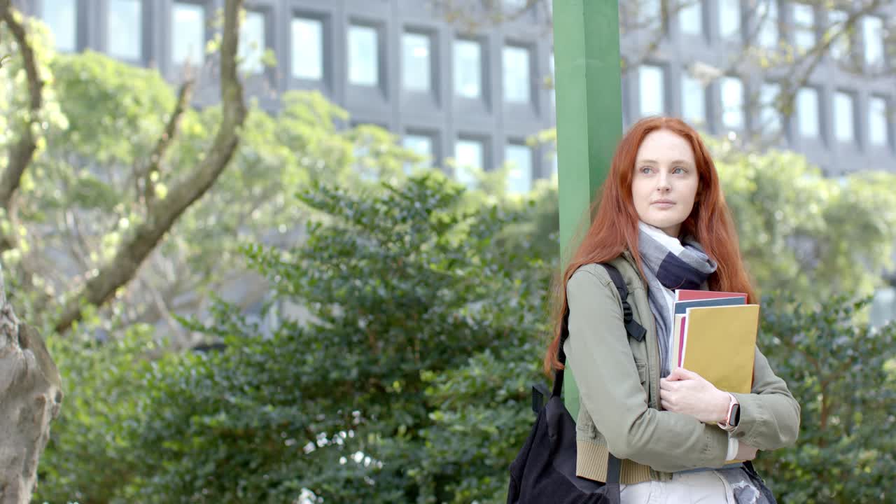 Donna appoggiata a un palo verde che tiene dei quaderni in mano, guarda di lato e aspetta un'amica in un cortile, spazio per il testo