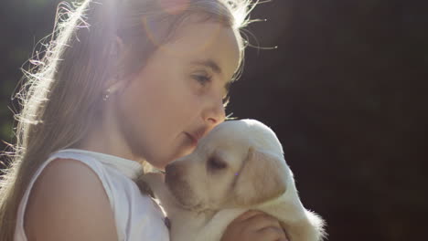 close-up view of a caucasian little girl holding and kissing a small labrador puppy in the park on a summer day