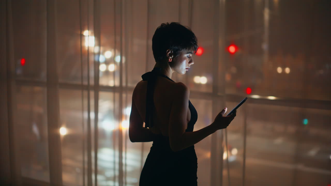Silhouette di donna che guarda la finestra controllando le notifiche del telefono di notte, vista da dietro.