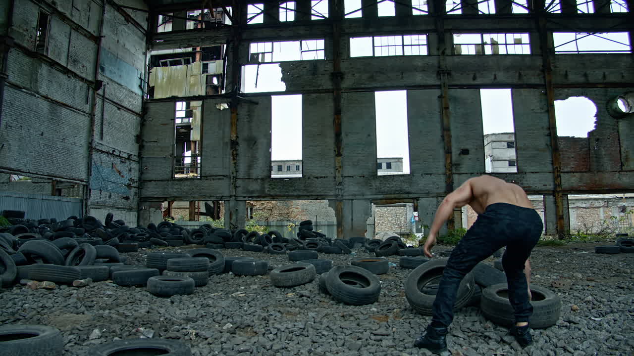 Shirtless man training in ruined plant. Strong athlete throwing old car tires while doing his workout in abandoned place. Cross fitness routine.